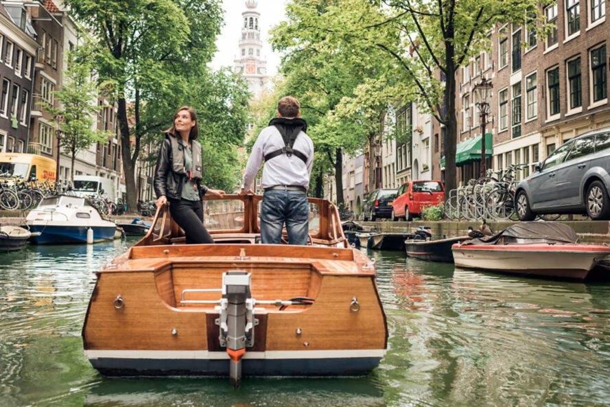 a wooden boat on a canal/lake area with trees lining the streets on either side of the canal. two people stand on the boat, with one person looking off into the distance