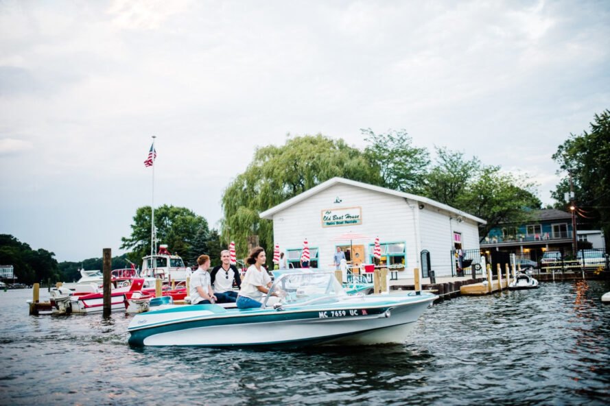 a boat on a lake with a white building on a dock behind it. three people are on the boat, with one of them steering the boat